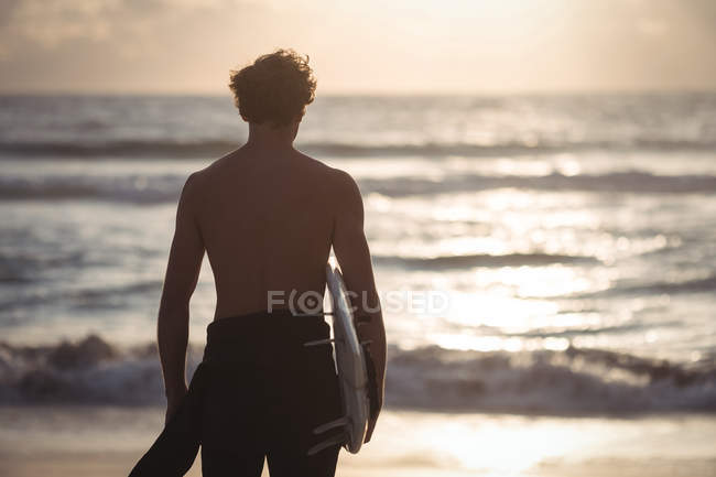 Вид сзади на человека, несущего доску для серфинга, стоящего на пляже в сумерках — стоковое фото