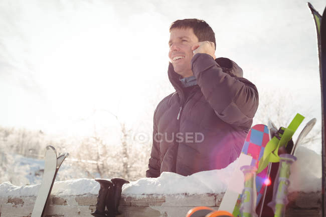 Улыбающийся мужчина в зимней одежде разговаривает по телефону возле заснеженного забора — стоковое фото