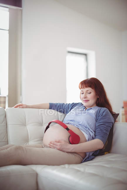 Вагітна жінка з навушниками на животі розслабляється на дивані у вітальні — стокове фото