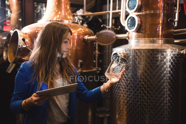 Frau mit digitalem Tablet untersucht Flüssigkeit im Becher in Bierfabrik — Stockfoto
