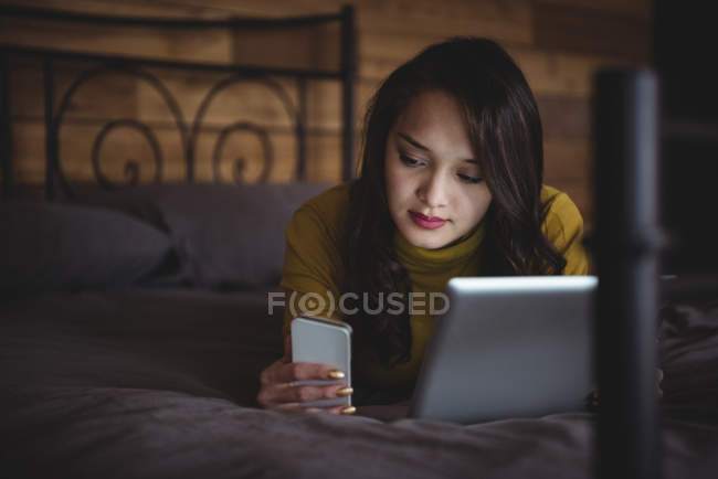 Женщина лежит на кровати с помощью цифрового планшета и мобильного телефона в спальне дома — стоковое фото