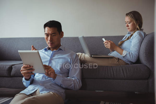 Couple utilisant téléphone portable, ordinateur portable et tablette numérique dans le salon à la maison — Photo de stock