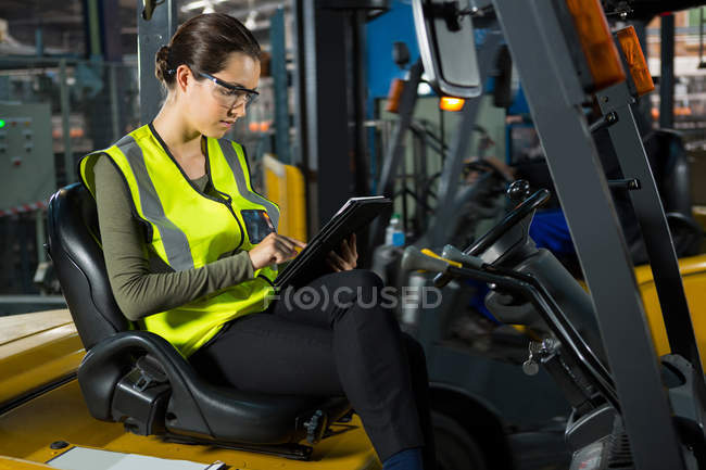 Confident female worker using digital tablet while sitting on forklift in warehouse — Stock Photo