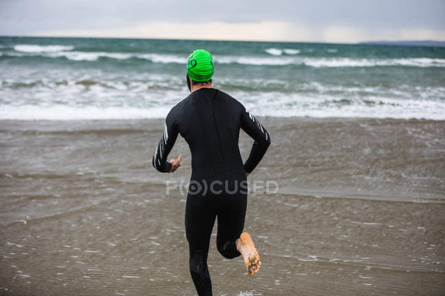 Vista traseira do atleta em terno molhado correndo na praia — Fotografia de Stock