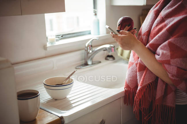 Середина жінки, використовуючи мобільний телефон, тримаючи яблуко на кухні вдома — стокове фото
