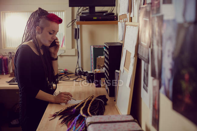 Женщина-парикмахер разговаривает по мобильному телефону и делает заметки в магазине дредов — стоковое фото