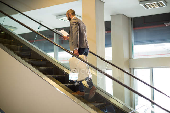 Вид сзади бизнесмена, стоящего на эскалаторе с багажом в аэропорту — стоковое фото
