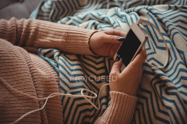 Mujer sentada en el sofá escuchando música en el teléfono móvil en la sala de estar en casa - foto de stock