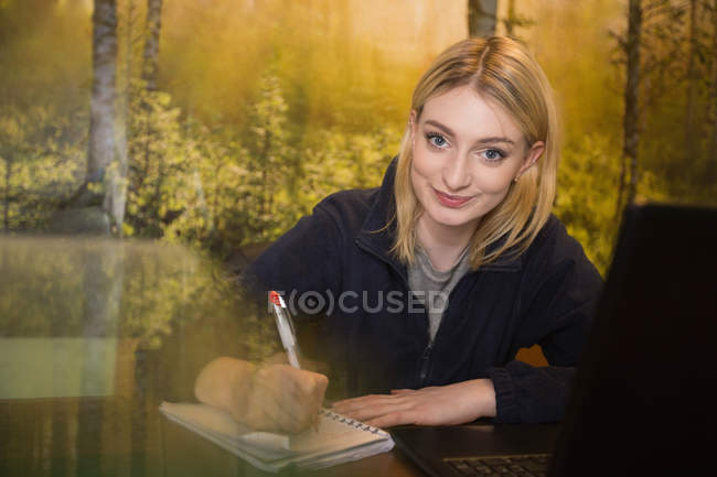 Щаслива жінка сидить за столом з ноутбуком і пише на блокноті в офісі — стокове фото