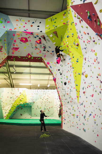 Formateur aidant femme tout en grimpant sur un mur artificiel dans la salle de gym — Photo de stock
