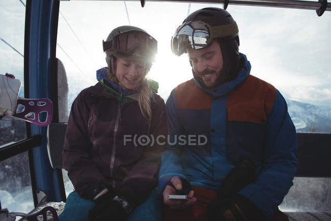 Happy couple using mobile phone while sitting in overhead cable car against sky during winter — Stock Photo