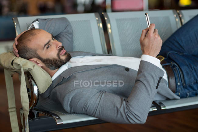 Empresário usando telefone celular enquanto deitado em cadeiras na área de espera no terminal do aeroporto — Fotografia de Stock