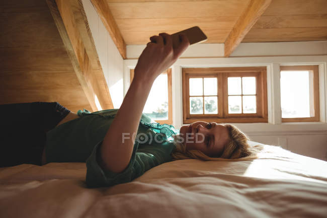 Belle femme couchée sur le lit et utilisant un téléphone portable dans la chambre à coucher à la maison — Photo de stock