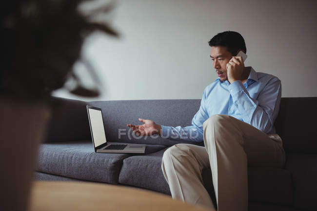 Чоловік розмовляє на мобільному телефоні, використовуючи ноутбук у вітальні вдома — стокове фото