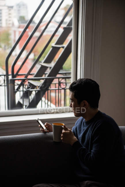 Homem usando telefone celular enquanto toma uma xícara de café em casa — Fotografia de Stock