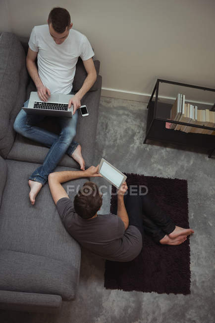 Couple gay utilisant tablette numérique et ordinateur portable dans le salon à la maison — Photo de stock