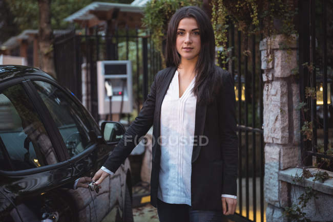 Vista frontal da bela mulher de pé perto do carro na estação de carregamento do veículo elétrico — Fotografia de Stock