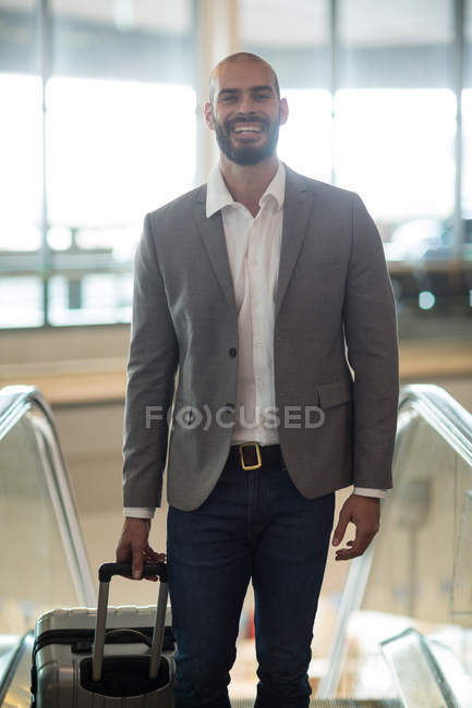 Hombre de negocios sonriente parado con el equipaje en la escalera mecánica en la terminal del aeropuerto - foto de stock