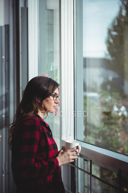 Mulher atenciosa olhando pela janela enquanto toma café no escritório — Fotografia de Stock