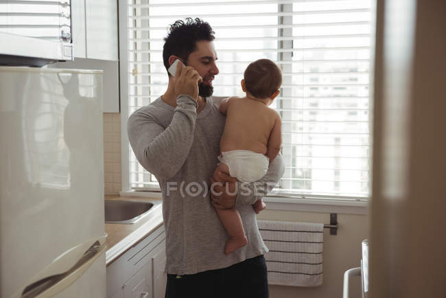 Батько розмовляє по мобільному телефону, тримаючи дитину на кухні — стокове фото