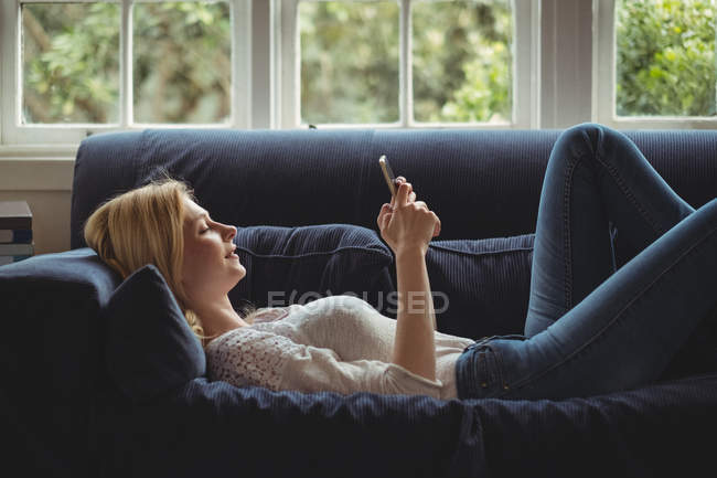 Hermosa mujer acostada en el sofá y el uso de teléfono móvil en la sala de estar en casa - foto de stock