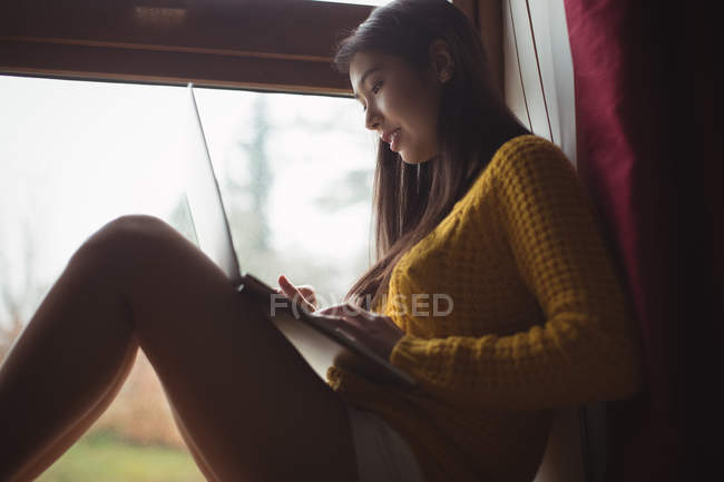 Mujer usando el ordenador portátil mientras está sentado en el alféizar de la ventana en casa - foto de stock