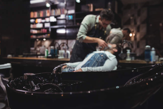Primer plano del lavabo con cliente de afeitado de peluquero en segundo plano en la peluquería - foto de stock