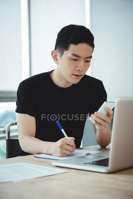 Business executive writing on diary while using mobile phone in office — Stock Photo