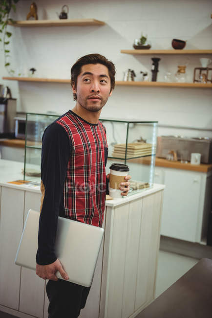 Homme tenant un ordinateur portable et une tasse de café jetable dans un café — Photo de stock