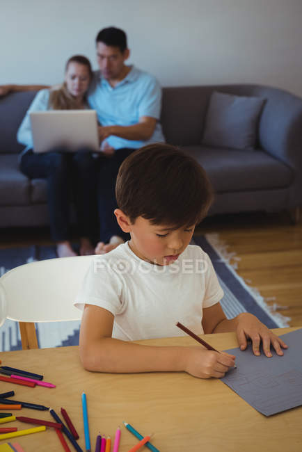Уважний хлопчик малює в папері, а його батьки використовують ноутбук у фоновому режимі вдома — стокове фото