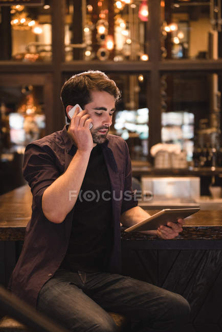 Hombre hablando en el teléfono móvil mientras usa la tableta digital en el mostrador de bar - foto de stock