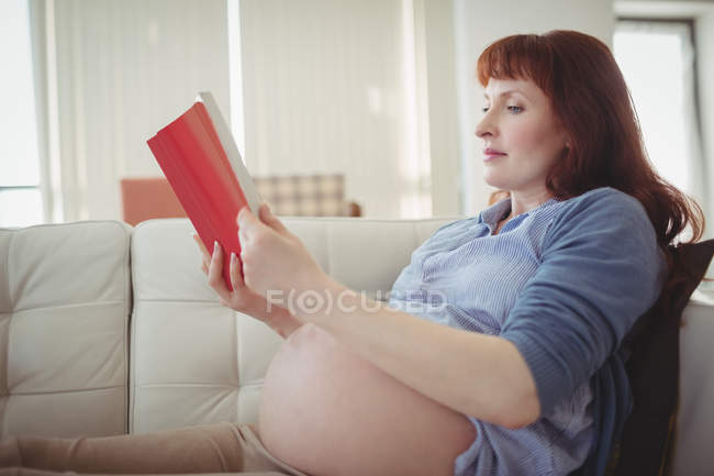 Mujer embarazada leyendo libro en el sofá en la sala de estar en casa - foto de stock
