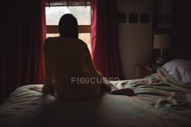 Woman sitting on bed and looking through window at home, rear view — Stock Photo