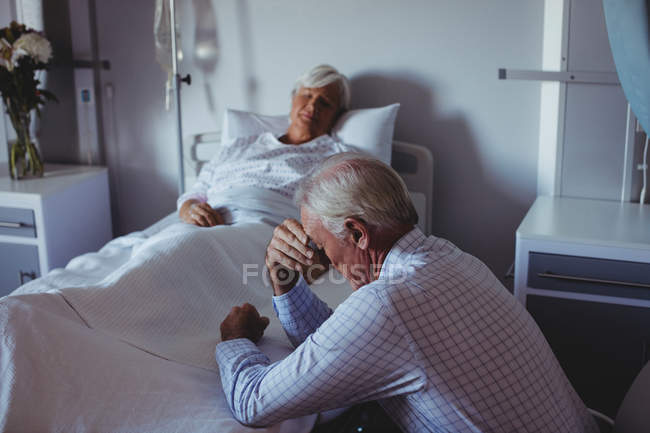 Нездоровая женщина спит на кровати, а обеспокоенный мужчина сидит рядом с ее кроватью в больнице — стоковое фото