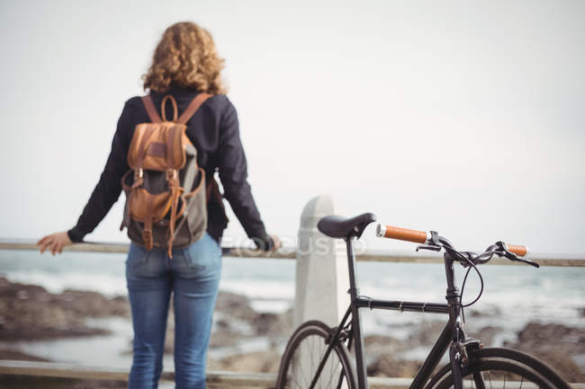 Vista trasera de una mujer de pie con la bicicleta cerca de la orilla del mar - foto de stock