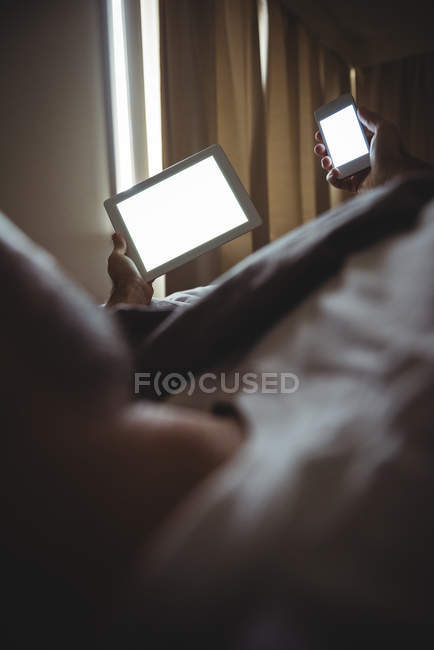 Вид сзади на человека, который использует свой мобильный телефон и цифровой планшет, отдыхая на кровати в спальне — стоковое фото