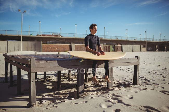 Pensiero surfista seduto su una piattaforma di legno con tavola da surf sulla spiaggia — Foto stock