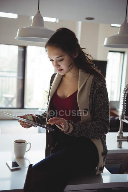 Mujer sentada en la encimera de la cocina usando tableta digital en casa - foto de stock