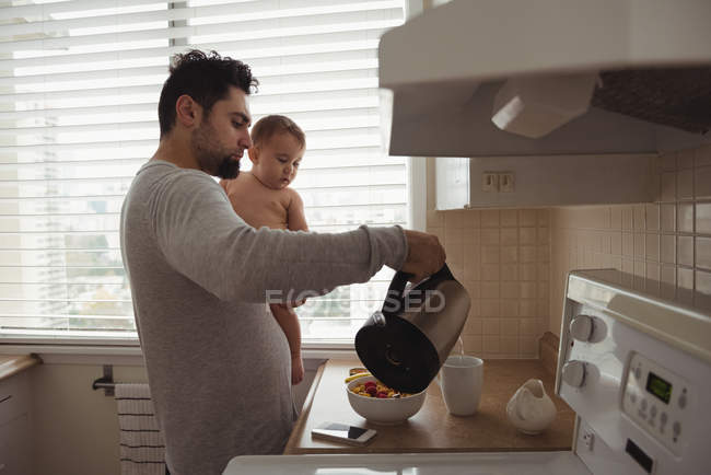 Padre preparando el desayuno mientras sostiene al bebé en la cocina - foto de stock