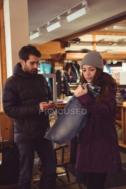 Frau wählt Kleidung in einem Bekleidungsgeschäft aus, während Mann Handy benutzt — Stockfoto