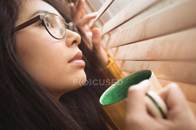 Mulher olhando pela janela enquanto toma café em casa — Fotografia de Stock