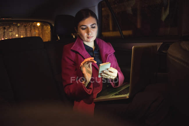 Geschäftsfrau sitzt mit Laptop im Auto und benutzt Handy — Stockfoto