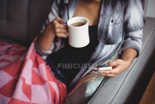 Femme utilisant un téléphone portable tout en tenant une tasse de café dans le salon à la maison — Photo de stock
