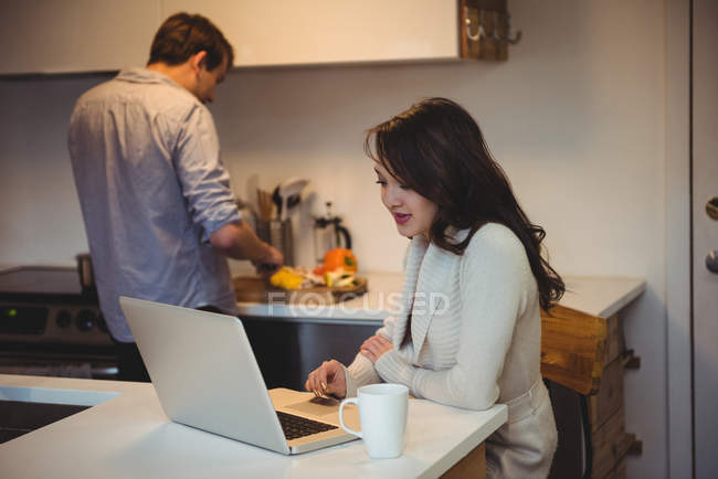 Frau benutzt Laptop, während Mann im Hintergrund in Küche arbeitet — Stockfoto