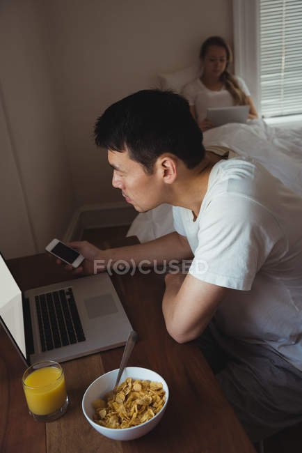 Чоловік використовує мобільний телефон і ноутбук під час сніданку в спальні вдома — стокове фото