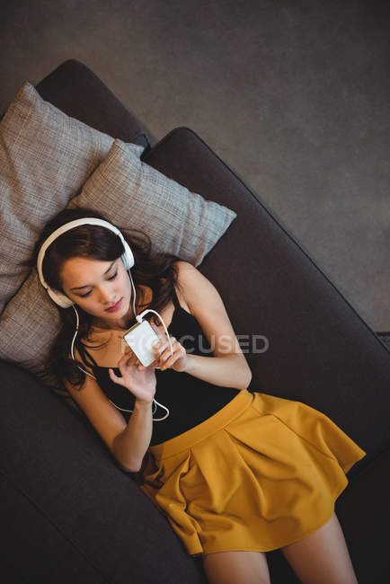 Женщина лежит на диване, слушая музыку с мобильного телефона дома — стоковое фото