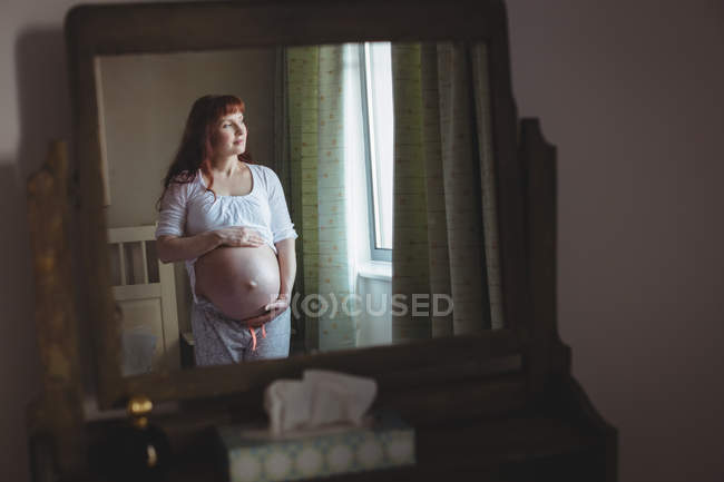 Reflection of pregnant woman looking through window in bedroom at home — Stock Photo