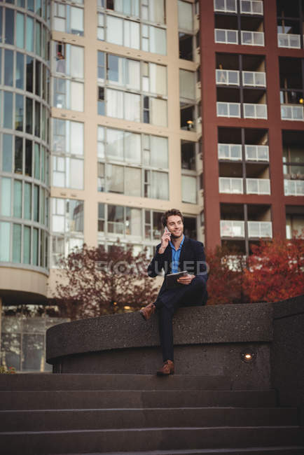 Homme d'affaires parlant sur un téléphone portable et tenant une tablette numérique près d'un immeuble de bureaux — Photo de stock