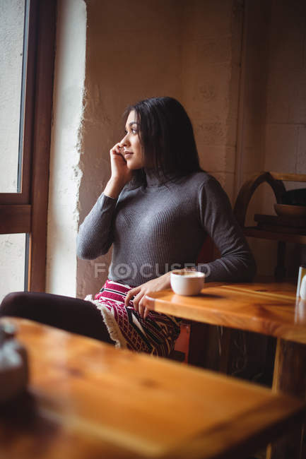 Mujer hablando por teléfono móvil mientras toma una taza de café en la cafetería - foto de stock