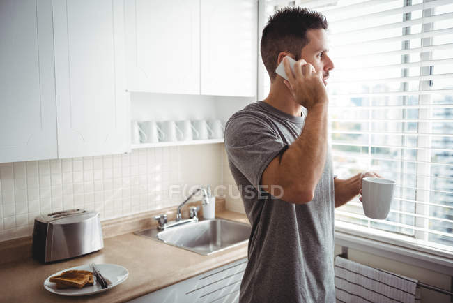 Мужчина разговаривает по мобильному телефону за чашечкой кофе дома на кухне — стоковое фото